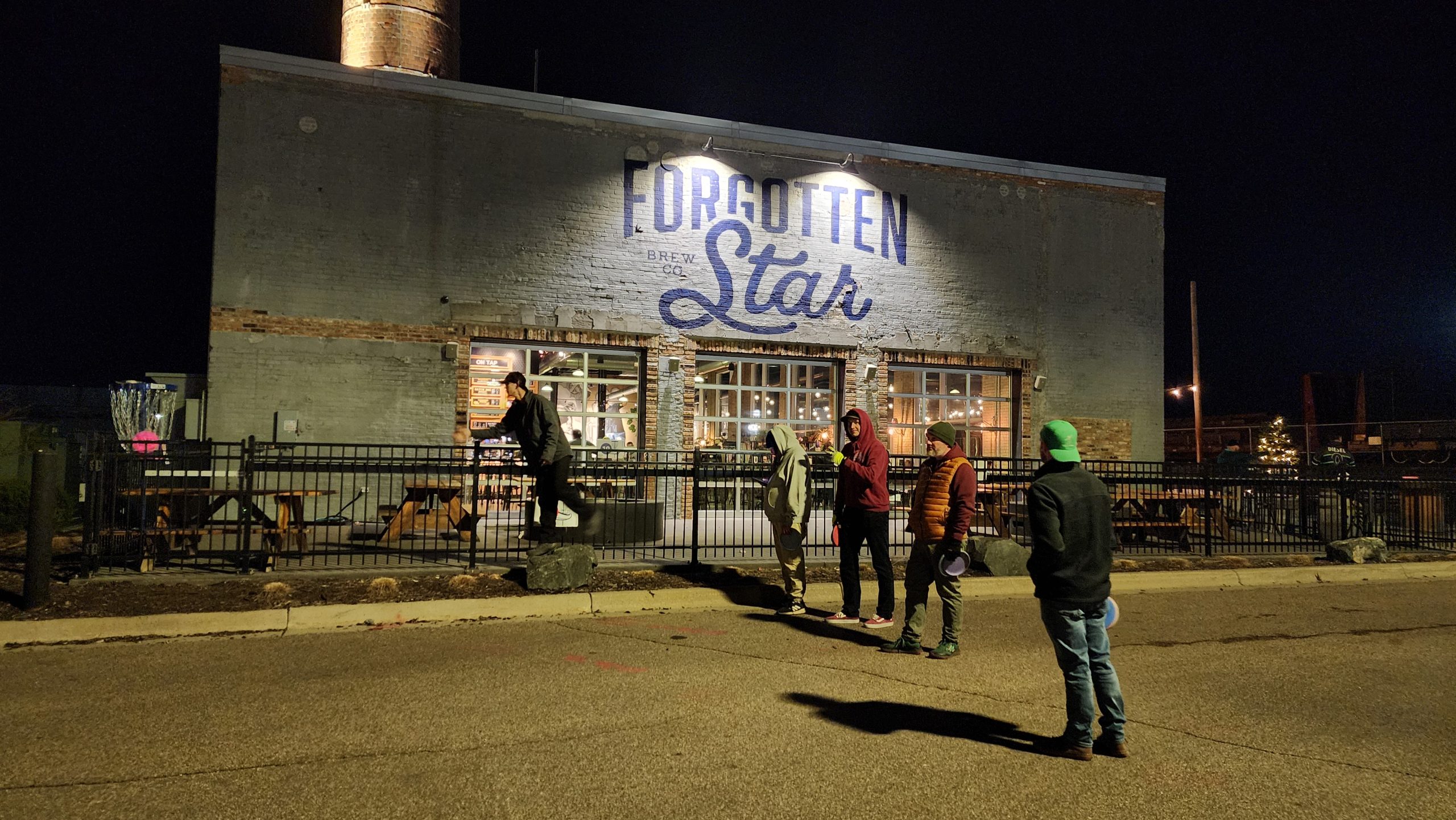 Players putting toward a disc golf basket outside Forgotten Star Brewing at night.